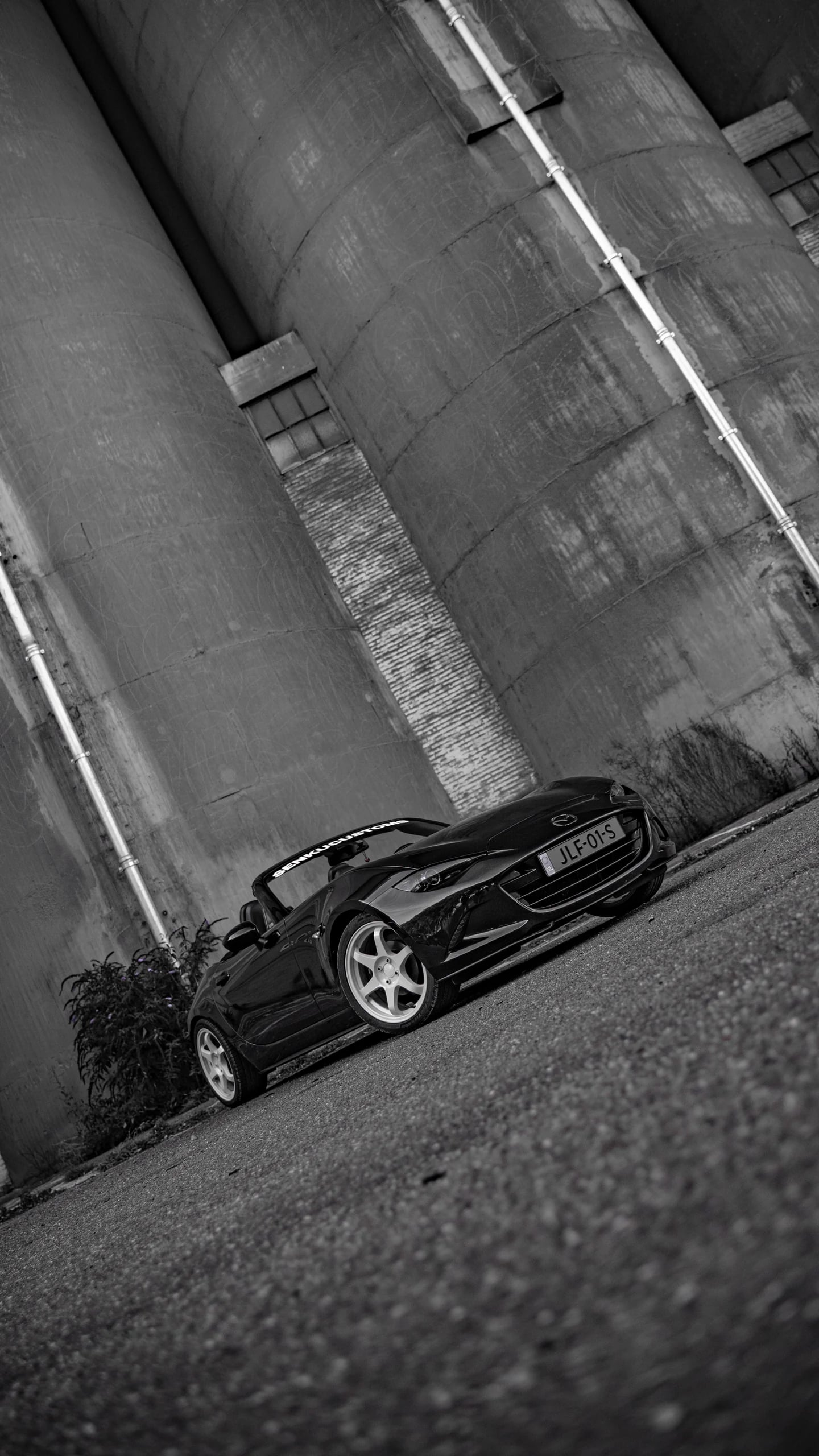 Person in an oni-print hoodie and grey joggers between a black MX-5 convertible and MX-5 RF by industrial silos
