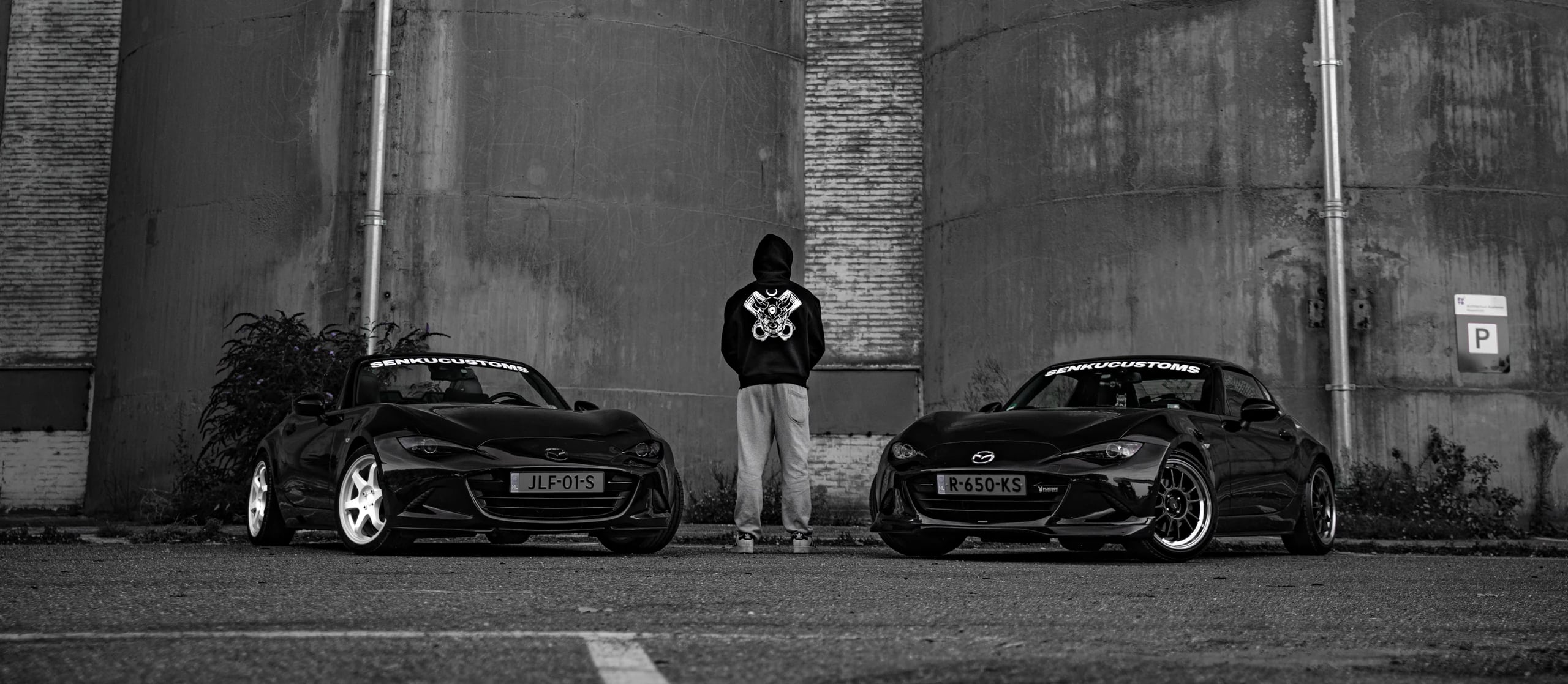 Hooded figure in an oni-print hoodie standing between two black Mazda MX-5s in front of concrete silos