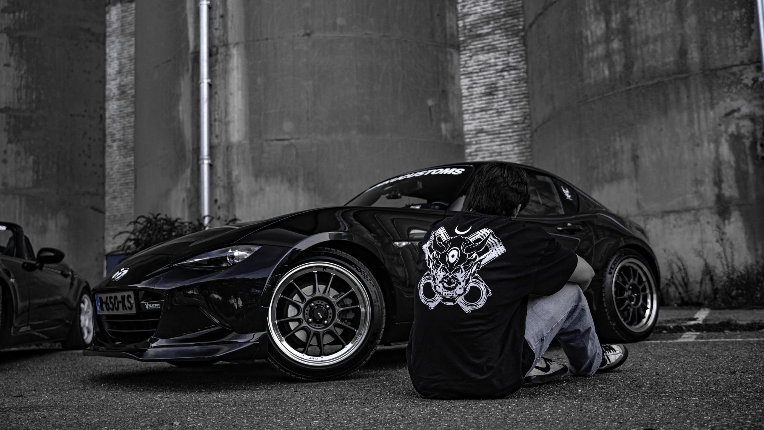 Person in an oni-print tee crouched beside a lowered black Mazda MX-5 RF on bronze-lipped wheels