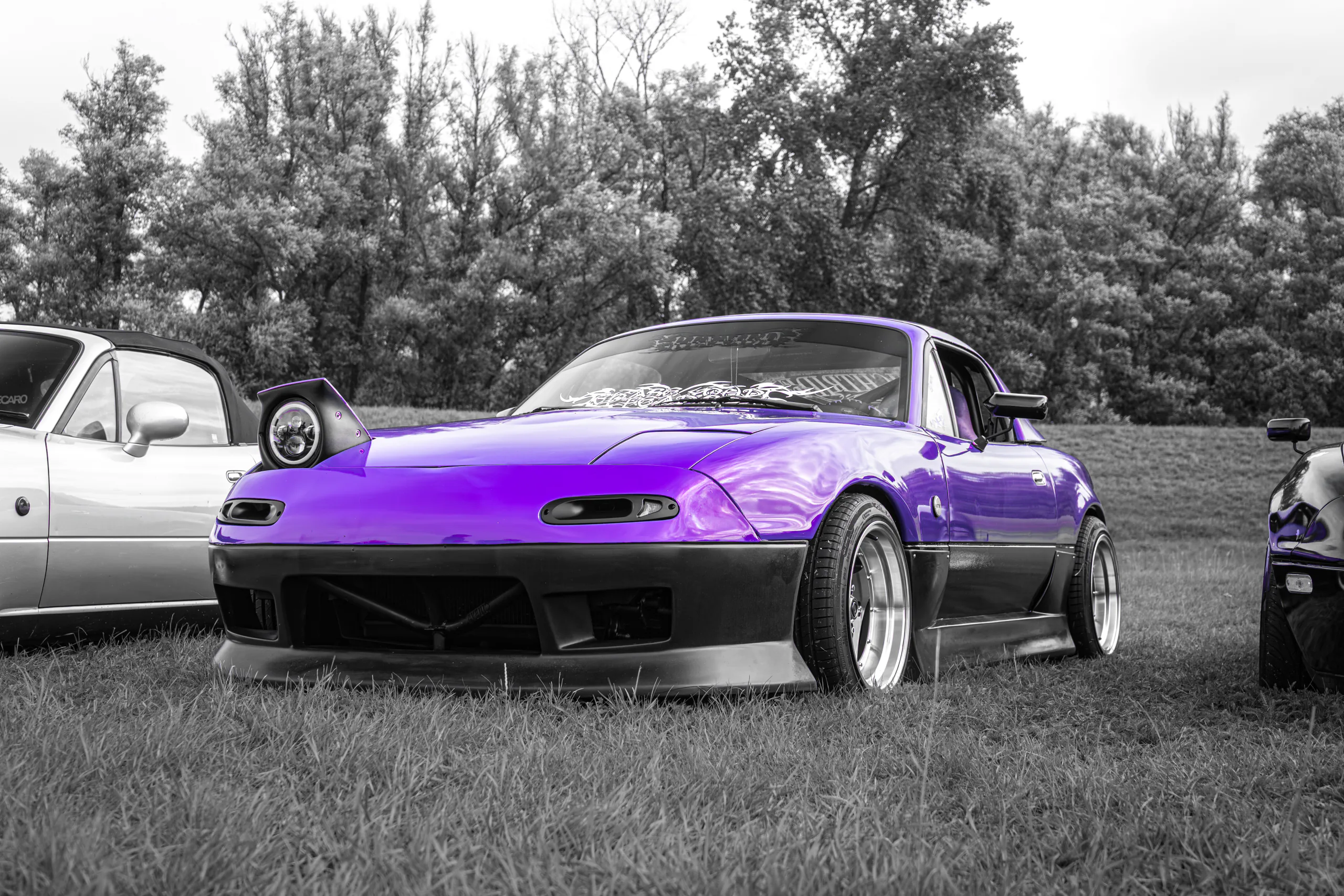 Purple Mazda MX-5 NA with one pop-up headlight raised, parked on the grass at a car meet, the rest of the scene in black and white